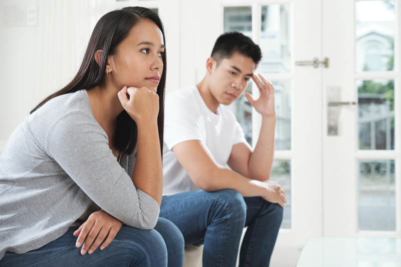 An Asian couple sit apart looking to be in tense silence. Person further from the viewer has short black hair and looks pensive, with one hand by their temple.  They are wearing a white shirt and blue jeans.  The person sitting closer to the audience has long dark brown hair, looking pensively with a hand on their chin.  They are wearing a grey top and jeans.  In the background are a white wall and a glass door. 