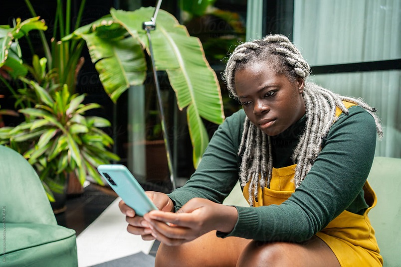 A Black person with silver dreadlocks to their chest is sitting down and looking worried at their mint- coloured cell phone in their hands.  They are wearing a deep green long sleeve top and yellow overalls.  They are sitting on a light green couch.  Behind them are muted green curtains and bright green leafy plants.  