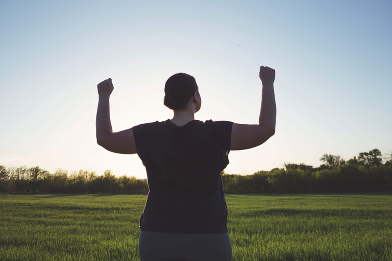 A fat person stands with their back to the viewer with raised arms looking confident.  They have their hair in a bun and is wearing dark shirt and pants.  They are facing an open green field with green trees and blue sky.  