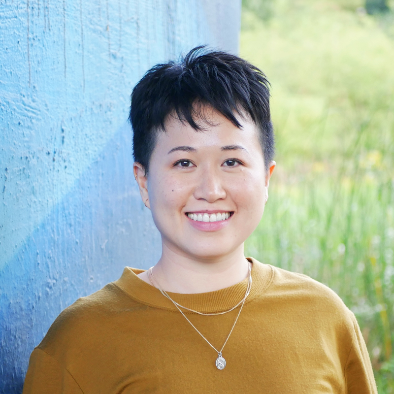 Jules is an East Asian person with short black hair and wearing a mustard coloured sweater with two gold necklaces. They are smiling and standing in front of a blue wall with greenery to the side.